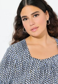 Blusa blu e bianca a pois con scollatura quadrata e dettagli arricciati, realizzata in un tessuto morbido. La modella indossa orecchini a cerchio in argento.