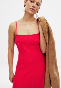 Röd figursydd klänning med spaghettiremmar, vertikala sömmar och en mjuk brun mockajacka hängande över ena armen. Stora hängande örhängen.