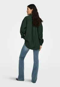 Dark green oversized shirt with long sleeves, relaxed fit, visible back seam, paired with light blue flared jeans and black shoes.