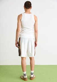 Débardeur blanc et shorts blancs avec des accents latéraux à motifs. Chaussures marron avec des chaussettes rayées blanches et noires. Vue de dos.