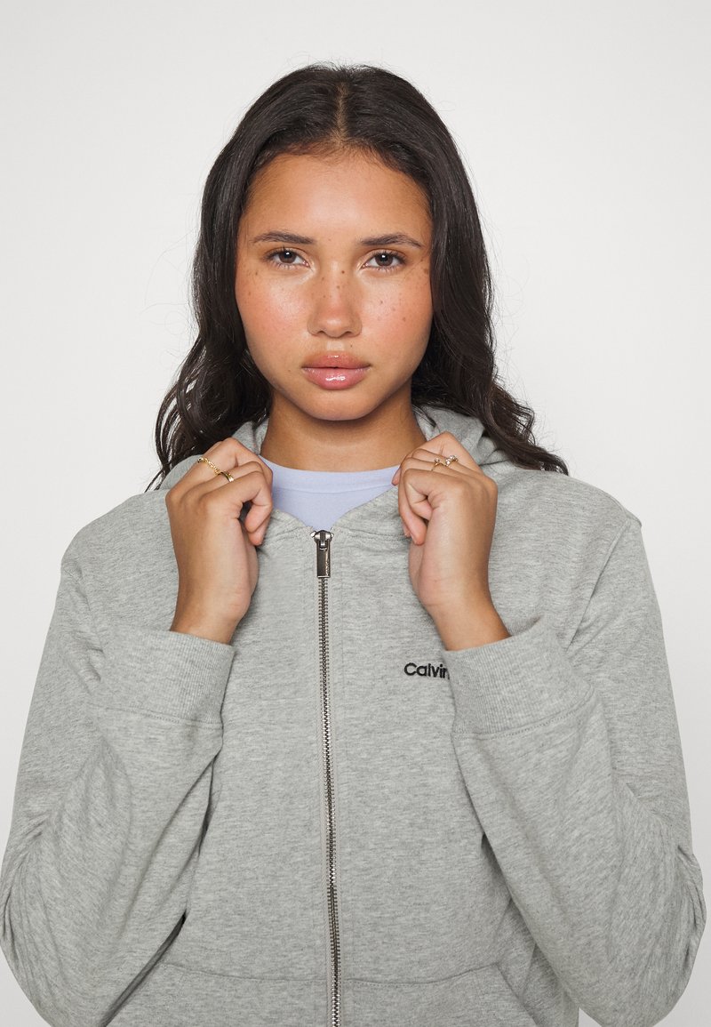 Sudadera gris con cremallera hecha de un tejido suave, con un cuello alto, puños acanalados y un pequeño logo negro en el pecho. El modelo sostiene la cremallera.