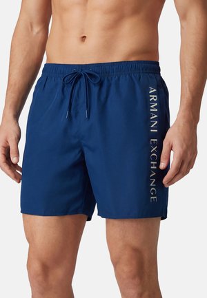 Torso maschile che indossa pantaloncini da bagno blu navy con elastico in vita e coulisse, con scritta verticale "Armani Exchange" sul lato destro.