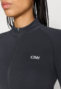 Melna sportiska zip-up jaka ar augstu apkakli. Ievērojumā ir sudraba logo uz krūtīm un gluda, elastīga auduma ar plakaniem šuvēm.