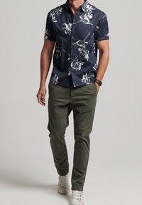 Camisa de manga corta azul marino con patrón floral blanco, combinada con pantalones verde oliva y zapatillas blancas, con un ajuste relajado y cierre de botones.