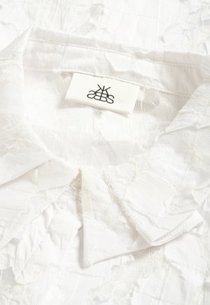 Camicia bianca con colletto in pizzo, motivo trapuntato e un'etichetta in tessuto con un logo nero simmetrico cucita all'interno della scollatura.