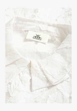 Cămașă albă cu guler din dantelă, având un model matlasat și o etichetă din material cu un logo negru simetric cusută în interiorul gâtului.