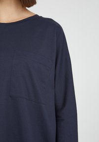 Tummansininen pitkähihaisellinen paita, joka on valmistettu pehmeästä puuvillasta. Paidassa on neliömäinen tasku vasemmassa rinnassa ja pyöreä kaula-aukko.