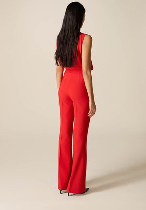 Vrouw met lang donker haar, gekleed in een mouwloos rood topje en bijpassende wijde rode broek, staand tegen een beige achtergrond.