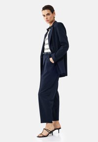 Vrouw in een marineblauwe blazer en broek met een gestreept top, zwarte riem, gouden oorbellen en zwarte hakken, staand met één hand in de zak.