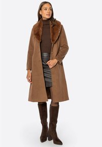 Brązowy wełniany płaszcz z kołnierzem ze sztucznego futra, długimi rękawami, zapięciem na guziki oraz długością do kolan, zestawiony z ciemną skórzaną spódnicą i brązowymi kozakami.