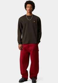 Camicia a maniche lunghe marrone scuro con un piccolo logo, abbinata a pantaloni rossi larghi e scarpe nere, caratterizzata da un design e una texture semplici.