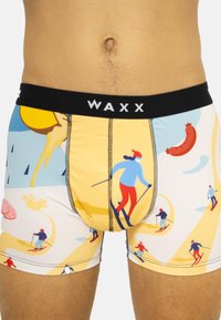 Caleçons de boxe avec un fond jaune présentant des motifs de ski, une taille noire avec "WAXX" et des coutures contrastées.
