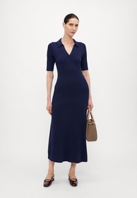 Robe en tricot bleu marine à manches courtes, décolleté en V, texture côtelée et silhouette ajustée, associée à un sac à main marron et des chaussures foncées.