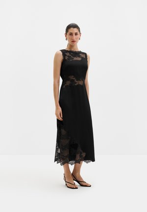 Femme debout portant une robe noire sans manches avec des panneaux en dentelle et un ourlet asymétrique, associée à des sandales noires à brides.