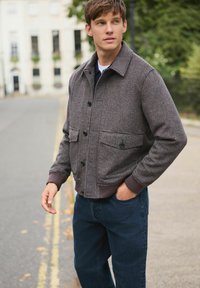 Young man wearing a brown checkered jacket and dark jeans standing on a street, looking to the side with one hand in his pocket.