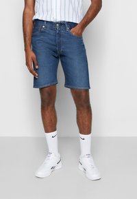 Shorts de mezclilla, azul oscuro, hasta la rodilla. Llevados con zapatillas deportivas blancas y una camiseta a rayas. Con bolsillos frontales y diseño de corte recto.