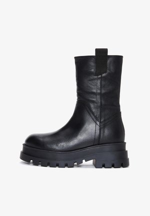 Botín de cuero negro con forma afilada, suela gruesa de goma y una lengüeta en la parte trasera. Presenta detalles cosidos y una textura suave.