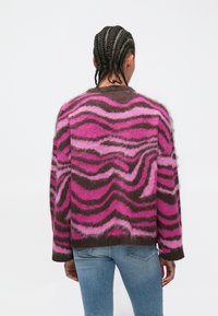 Pessoa com cabelo trançado, vestindo um suéter ondulado rosa e castanho e jeans azul claro, em pé de frente para um fundo branco.