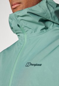 Chaqueta impermeable verde claro con cuello alto, cierre de cremallera y logotipo bordado en negro de "berghaus" en el pecho. Textura suave.