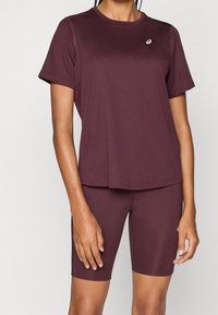 Person som bär en mörklila kortärmad sporttröja och matchande åtsittande cykelbyxor, stående mot en neutral bakgrund.