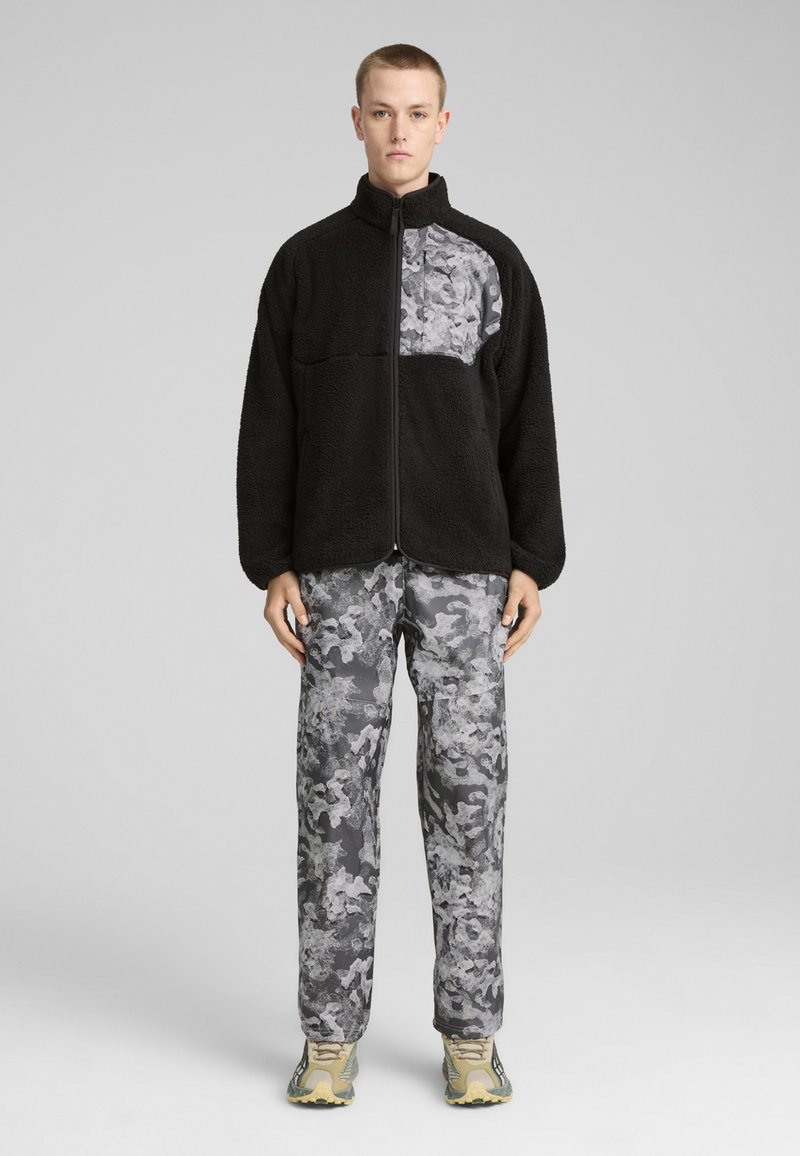 Must mustriga jope, millel on hall mustriga paneel, koos hallide camo pükstega ja tan värvi spordijalatsitega, luues ühtse outfitsi.