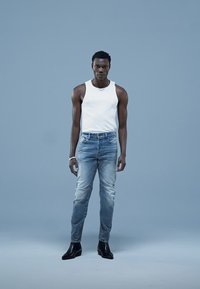 Vit ärmlös linne, ljusblå slim-fit jeans med blekta partier, svarta ankelboots, stående mot en enfärgad blå bakgrund.