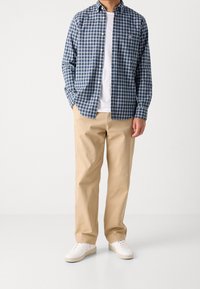 Mężczyzna w beżowych spodniach, białych trampkach, białej koszulce i niebieskiej koszuli w kratę z jednym ręką w kieszeni.