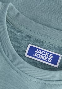 Γιακάς από λεπτομέρεια βαμβακερής μπλούζας σε ανοιχτό γαλάζιο με ριμπ υφή. Η ετικέτα γράφει «JACK & JONES» σε λευκό χρώμα σε μπλε φόντο. Λείο ύφασμα.