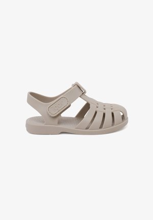 Sandal infantil de color beige claro, hecho de plástico flexible. Presenta un diseño descubierto en la parte delantera, strap ajustable y una plantilla texturizada elevada para mayor agarre.