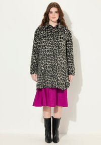 Cappotto maculato con bottoni neri, maniche lunghe e vestibilità rilassata, indossato sopra un vivace vestito rosa. La modella indossa anche stivali neri fino al ginocchio.