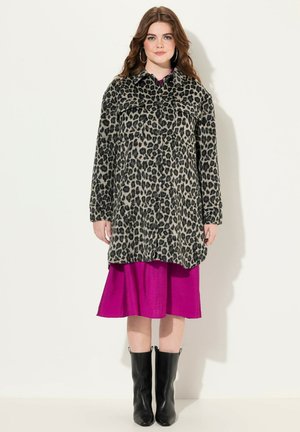Cappotto maculato con bottoni neri, maniche lunghe e vestibilità rilassata, indossato sopra un vivace vestito rosa. La modella indossa anche stivali neri fino al ginocchio.