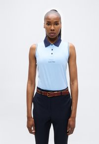 Γυναίκα που στέκεται όρθια φορώντας ανοιχτό μπλε αμάνικο πόλο με μαύρο γιακά και σκούρο παντελόνι με καφέ ζώνη, μπροστά σε απλό φόντο.