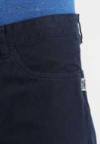 Gros plan sur un pantalon Tommy Jeans bleu marine montrant la poche avant, la boucle de ceinture et l'étiquette sur la couture latérale.