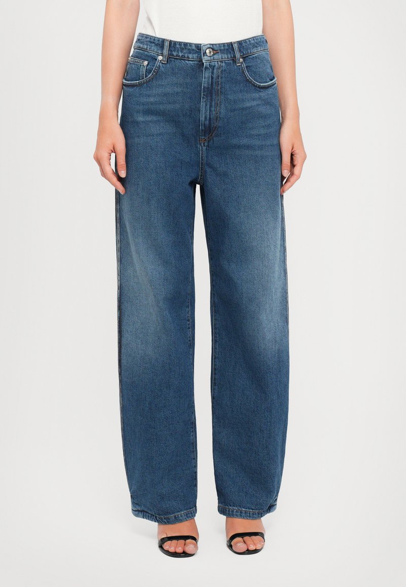 Femme portant un jean large taille haute bleu et des escarpins ouverts noirs, se tenant devant un fond blanc uni.