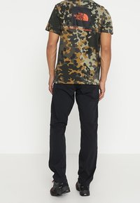 T-shirt à manches courtes avec motif camouflage en vert, marron et noir ; arbore un logo carré dans le dos. Porté avec un pantalon et des chaussures noires.