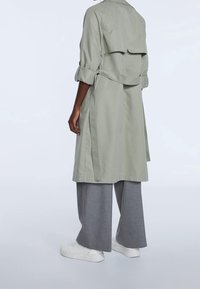 Trenca verde claro con cintura ceñida, mangas enrolladas y un diseño de espalda en capas, combinada con pantalones de pierna ancha grises y zapatillas blancas.