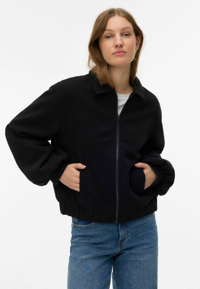 Veste noire à fermeture éclair avec col, manches surdimensionnées et poignets élastiques, associée à un jean droit bleu clair. Texture de tissu lisse.