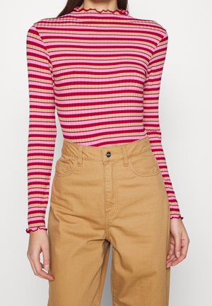 Woman wearing ribbed, long-sleeve striped top in red, pink, and beige with ruffled edges, paired with high-waisted tan pants.