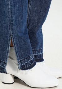 Botines de cuero blanco hasta el tobillo con punta afilada y tacones gruesos, combinados con jeans de mezclilla oscura de corte deshilachado y con aberturas laterales.