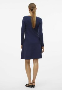 Mörkblå långärmad klänning med utsvängd fåll, stickad textur och rund halsringning, bärs med svarta sandaletter med öppen tå och klack.