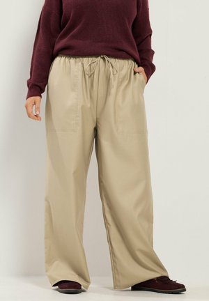 Personne portant un pantalon beige à jambes larges avec une taille élastique à cordon, associé à un pull bordeaux et des chaussures bordeaux assorties.
