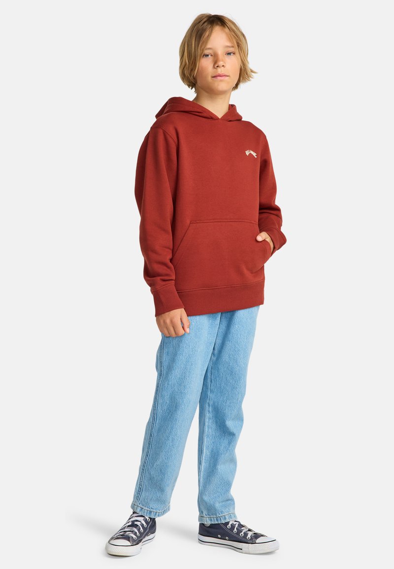 Felpa rossa con cappuccio, dotata di tasca frontale e dettaglio del logo, abbinata a pantaloni in denim blu chiaro e scarpe da ginnastica blu scuro.