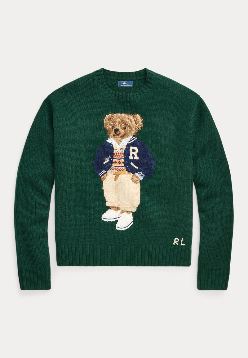 Pull vert foncé avec un ours en peluche tricoté portant une veste universitaire bleue, un pull à motifs, un pantalon beige et des chaussures blanches sur le devant.