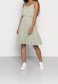 Ljusgrön, knälång klänning med en figurnära överdel, plisserad kjol och tunna spagettistilar, i kombination med vita sneakers.