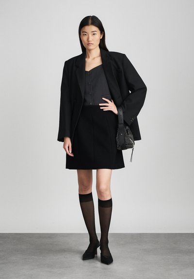 Črn blazer nad sivo majico, v kombinaciji s črno krilo. Model nosi prozorne črne do kolen nogavice in konične črne čevlje, drži biserno torbico.