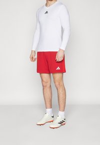 Camisa deportiva de manga larga blanca con el logo negro de Adidas, combinada con pantalones cortos rojos que tienen el logo blanco de Adidas, y botas de fútbol blancas con detalles negros.