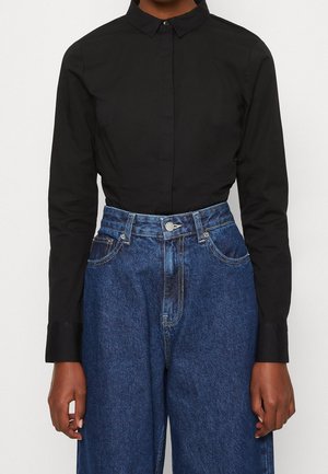 Person wearing a buttoned black long-sleeve shirt tucked into high-waisted blue denim jeans, standing against a plain background.