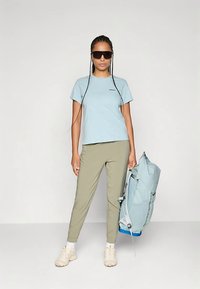 Ανοιχτό μπλε κοντομάνικο μπλουζάκι, ελαιδί στενά παντελόνια, μπεζ αθλητικά παπούτσια και ανοιχτό μπλε σακίδιο πλάτης κρατημένο στο ένα χέρι.