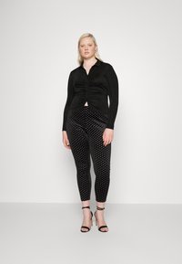 Camisa preta de manga comprida com franzido na frente, combinada com leggings pretas de veludo com bolinhas brancas. Modelo usa sandálias altas pretas.