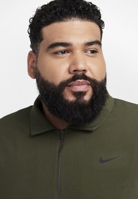 Vyras su garbanotais plaukais ir pilna barzda, vilkintis alyvuogių žalumo zip-up striukę su mažu juodu Nike logotipu, žiūri į dešinę.